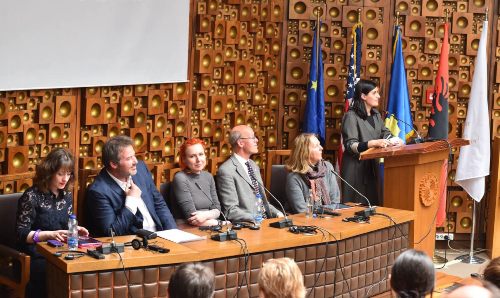 A panel of five people sat with microphones and one lady standing speaking 